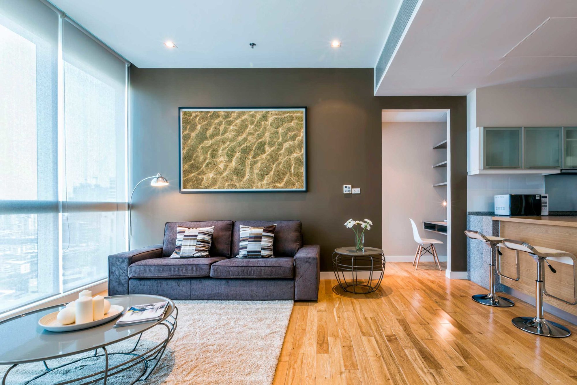 Wohnzimmer mit hellem Holzboden und grauer Couch vor einer grauen Wand mit großem Bild und einem Arbeitszimmer im Hintergrund